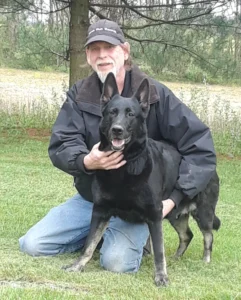 A man wearing a baseball cap kneeling beside his German Shepherd in a field of grass