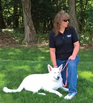 Betsy and Marley a white German Shepherd A woman with long brown hair, wearing blue jean, a dark blue polo shirt with white letters across the chest, and white sneakers is kneeling beside a white German Shepherd who is lay down in the green grass. The woman is wearing sunglasses as the sun is shining brightly on the pair. Trees are in the background.
